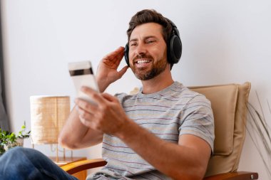 Kablosuz kulaklıkla müzik dinleyen, akıllı telefon tutan, evdeki koltukta oturan mutlu bir adam.
