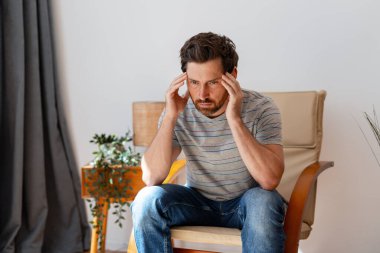 Evdeki sandalyede oturan bir adam, baş ağrısı ve stresten muzdaripken şakaklarına masaj yapıyor.