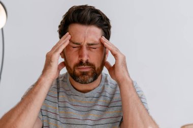 Sakallı, gözleri kapalı beyaz bir adam stres ve gerilimden kaynaklanan baş ağrısını hafifletmek için şakaklarına masaj yapıyor.