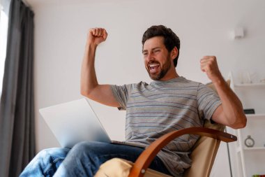 Mutlu erkek serbest çalışan bilgisayarında iyi haberler aldıktan sonra sevinçle kollarını kaldırıyor, ev işlerinden zevk alıyor.