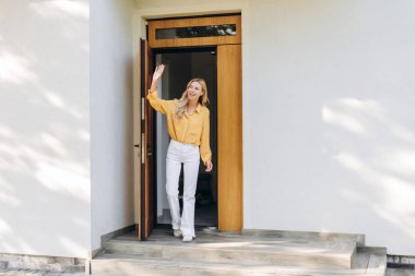 Gülümseyen kadın modern evden çıkarken el sallıyor, bir veda ve ikametgahından ayrılma hissini iletiyor, neşeli bir şekilde yeni başlangıçları kucaklıyor.