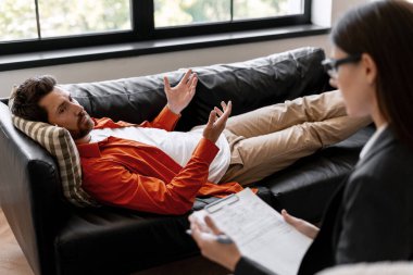 Adam kanepede uzanıyor, terapi sırasında aktif olarak konuşuyor ve el kol hareketi yapıyor ya da bir kadın psikoloğa danışmanlık yapıyor.