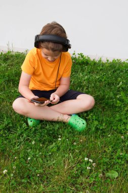 Çimlerin üzerinde oturan ve akıllı telefonu ve kulaklıkla en sevdiği müziği dinleyen bir çocuk. Beyaz arkaplan