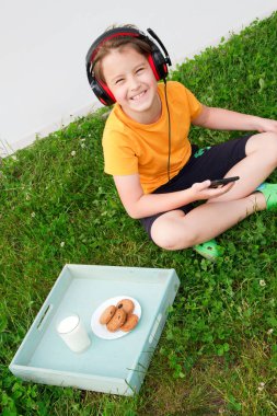 Çocuk çimlerin üzerinde oturuyor ve en sevdiği müziği dinliyor. Akıllı telefonunu ve kulaklığını kullanıyor. 