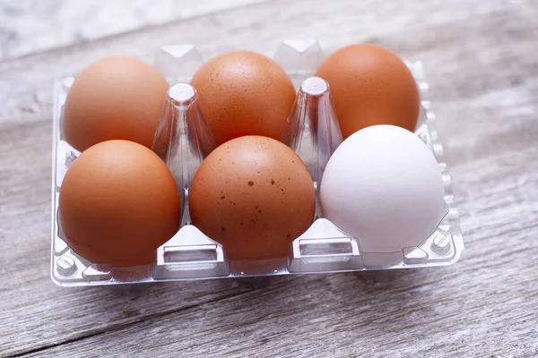 Six eggs plastic packing on wooden table. One different color of egg. Five brown and one white.