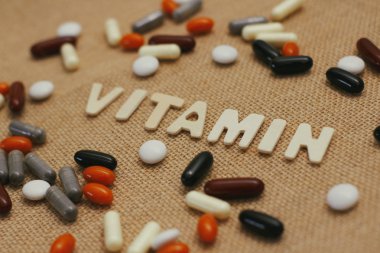 Kahverengi kaba kumaş doku üzerinde ahşap harflerle Vitamin kavram metni. Farklı renklerde hap ve kapsül seti. 