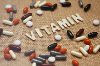 Kahverengi kaba kumaş doku üzerinde ahşap harflerle Vitamin kavram metni. Farklı renklerde hap ve kapsül seti. 