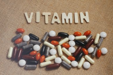 Kahverengi kaba kumaş doku üzerinde ahşap harflerle Vitamin kavram metni. Farklı renklerde hap ve kapsül seti. 