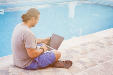 Diz üstü bilgisayarı olan genç adamlar havuz kenarında oturuyor. Açık hava iş yeri. Serbest meslek kavramı. İnternetten alışveriş. Tatilden işe.