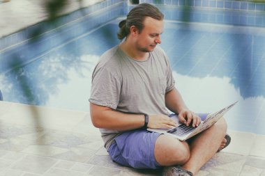 Diz üstü bilgisayarı olan genç adamlar havuz kenarında oturuyor. Açık hava iş yeri. Serbest meslek kavramı. İnternetten alışveriş. Tatilden işe.