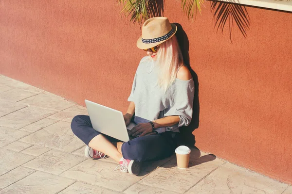 Dizüstü bilgisayarı, hasır şapkası, kırmızı ayakkabısı ve kağıttan kahvesi olan genç bir kadın serbest çalışmak ve online alışveriş için dışarıda oturuyor. Seyahat konsepti.