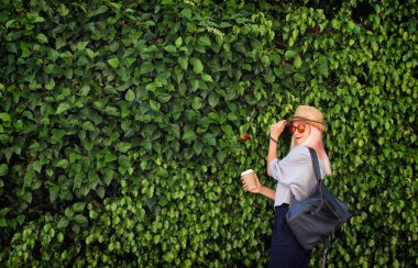 Kağıt bardak kahve, hasır şapka ve yeşil yapraklı sırt çantalı genç bir kadın. Pembe sarı saçlar. Güneş gözlüğü. Yaz zamanı..