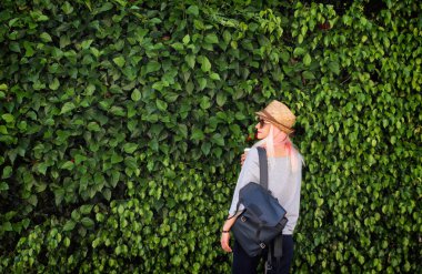 Kağıt bardak kahve, hasır şapka ve yeşil yapraklı sırt çantalı genç bir kadın. Pembe sarı saçlar. Güneş gözlüğü. Yaz zamanı..