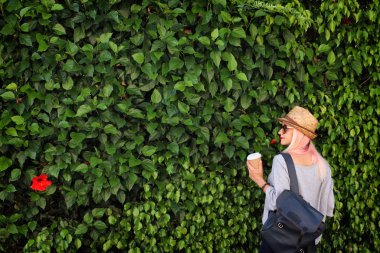 Kağıt bardak kahve, hasır şapka ve yeşil yapraklı sırt çantalı genç bir kadın. Pembe sarı saçlar. Güneş gözlüğü. Yaz zamanı..