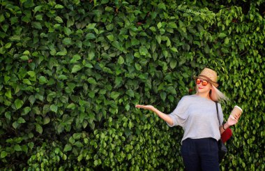 Kağıt kahve, güneş gözlüğü, hasır şapka, sarı saçlı ve deri sırt çantası olan genç kız. Yeşil yapraklar duvar arka plan. 