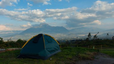 Bulutlu bir günde arka planda Imbabura volkanı olan bir çim tarlasında yeşil bir kamp çadırı.