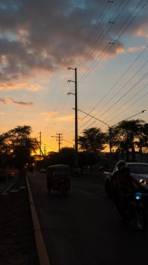 San Miguel de Piura, Piura / Peru - 5 Nisan 2019: Ağaçlar ve arabaların siyah siluetinde güneş ağacın arasında saklandığı bir caddede gün batımının görüntüsü