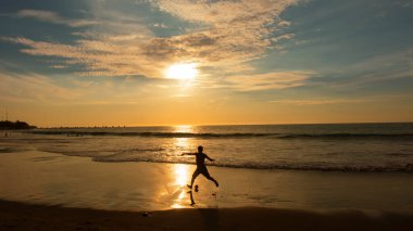 Genç adam gün batımında plajda futbol oynuyor. Güneş ışığı denize yansıyor.
