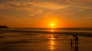 Baba ve kızı gün batımında kumsalda oynuyorlar. Güneş denizde parlıyor.