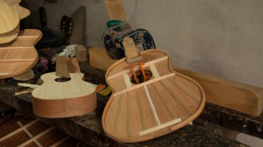 Marangozun tezgahındaki parçaları tutan baskınla birleştirilme sürecinde iki ahşap gitar ve arka planda gri duvar var.