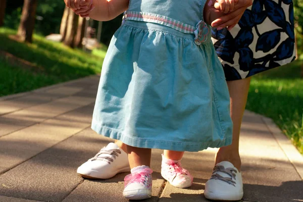 Mom teaches daughter to walk. Summer park, legs closeup. Mom with a one-year-old daughter in dresses are walking in the park. Teaching children, motherhood.