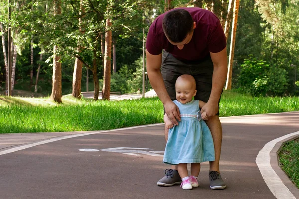 Dad teaches daughter to walk. Happy dad and daughter. Dad with a one-year-old daughter in a dress are walking in the park. Teaching children, fatherhood. The first steps.