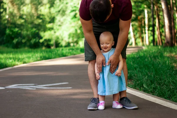 Dad teaches daughter to walk. Happy dad and daughter. Dad with a one-year-old daughter in a dress are walking in the park. Teaching children, fatherhood. The first steps.
