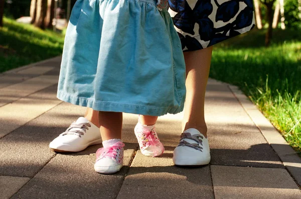 Mom teaches daughter to walk. Summer park, legs closeup. Mom with a one-year-old daughter in dresses are walking in the park. Teaching children, motherhood.