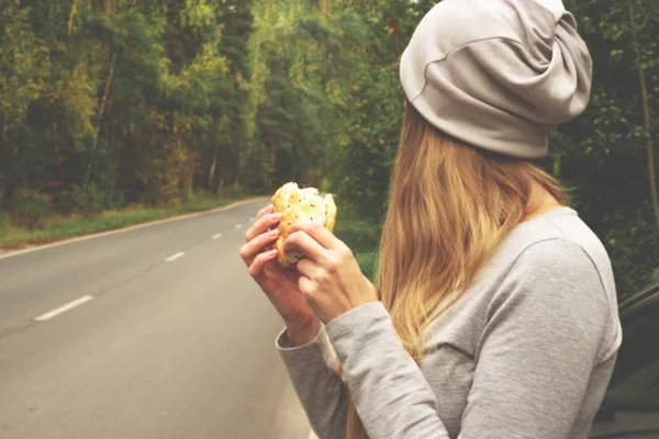 A young woman is eating a burger near a car on an empty road. Food on the trip. Food on the go. Autumn travel. Fast food.