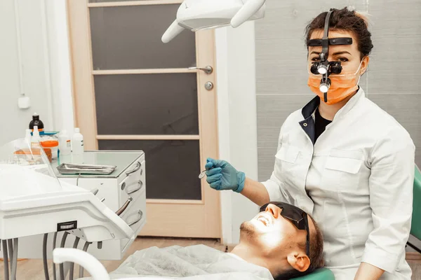 Dentist young woman treats a patient a man. The doctor uses disposable gloves, a mask and a hat. The dentist works in the patients mouth, uses a professional tool.