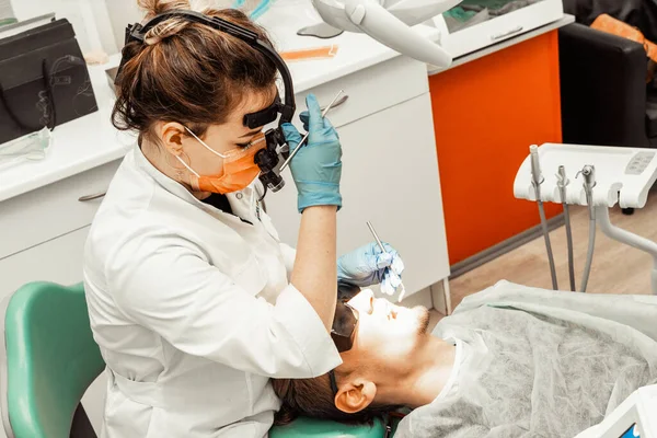 Dentist young woman treats a patient a man. The doctor uses disposable gloves, a mask and a hat. The dentist works in the patients mouth, uses a professional tool.