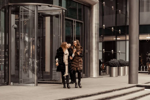 Female friendship, relationships. Two girls are drinking coffee on the street near glass office buildings, a corporation, a bank. The end of the working day of the manager. Coffee on the run.