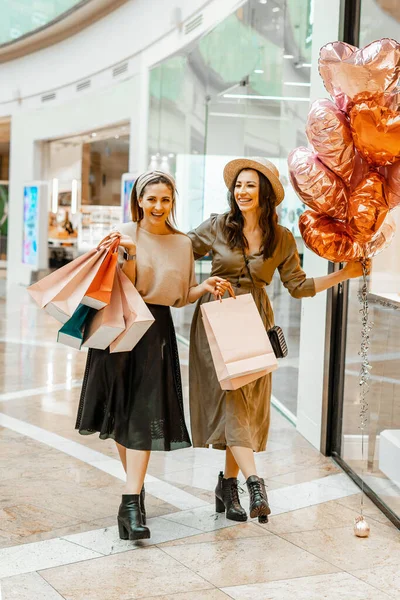 Shopping and entertainment, mall inside. Two beautiful girls with paper bags in the mall, heart-shaped balloons. The joy of consumption, Gift shopping, holiday.
