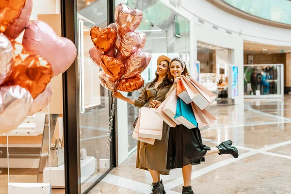 Shopping and entertainment, mall inside. Two beautiful girls with paper bags in the mall, heart-shaped balloons. The joy of consumption, Gift shopping, holiday.