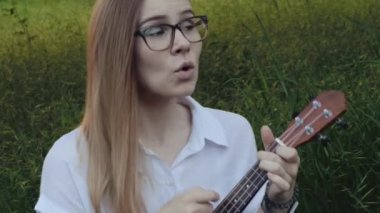 Ukulele çalan genç ve güzel bir kız. Gitar çalan beyaz bir kız. Genç güzel sarışın kadın gitar çalıyor ve çimlerin üzerinde otururken şarkı söylüyor. Kapat..