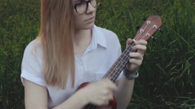 Ukulele çalan genç ve güzel bir kız. Gitar çalan beyaz bir kız. Genç güzel sarışın kadın gitar çalıyor ve çimlerin üzerinde otururken şarkı söylüyor. Kapat..