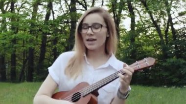 Ukulele çalan genç ve güzel bir kız. Gitar çalan beyaz bir kız. Genç güzel sarışın kadın gitar çalıyor ve çimlerin üzerinde otururken şarkı söylüyor. Kapat..