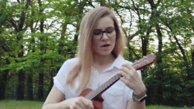 Ukulele çalan genç ve güzel bir kız. Gitar çalan beyaz bir kız. Genç güzel sarışın kadın gitar çalıyor ve çimlerin üzerinde otururken şarkı söylüyor. Kapat..