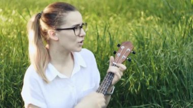 Ukulele çalan genç ve güzel bir kız. Gitar çalan beyaz bir kız. Genç güzel sarışın kadın gitar çalıyor ve çimenlerde otururken şarkı söylüyor..