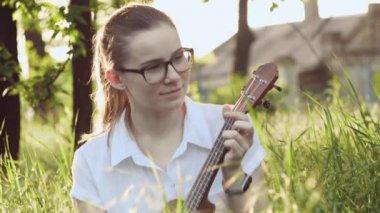 Ukulele çalan genç ve güzel bir kız. Gitar çalan komik beyaz kız. Genç ve güzel sarışın kadın gitar çalıyor çimlerin üzerinde oturuyor.