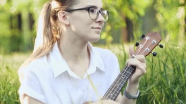 Ukulele çalan genç ve güzel bir kız. Gitar çalan komik beyaz kız. Genç ve güzel sarışın kadın gitar çalıyor çimlerin üzerinde oturuyor.
