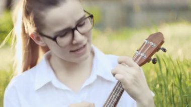 Ukulele çalan genç ve güzel bir kız. Gitar çalan beyaz bir kız. Genç güzel sarışın kadın gitar çalıyor ve çimlerin üzerinde otururken şarkı söylüyor. Kapat..