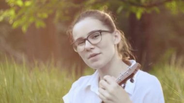 Ukulele çalan genç ve güzel bir kız. Gitar çalan beyaz bir kız. Genç güzel sarışın kadın gitar çalıyor ve çimlerin üzerinde otururken şarkı söylüyor. Kapat..