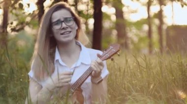 Ukulele çalan genç ve güzel bir kız. Gitar çalan beyaz bir kız. Genç güzel sarışın kadın gitar çalıyor ve çimlerin üzerinde otururken şarkı söylüyor. Kapat..