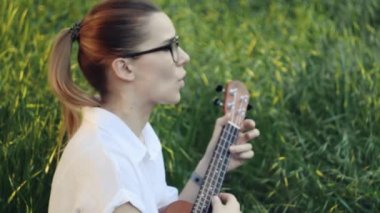 Ukulele çalan genç ve güzel bir kız. Gitar çalan beyaz bir kız. Genç güzel sarışın kadın gitar çalıyor ve çimlerin üzerinde otururken şarkı söylüyor. Kapat..