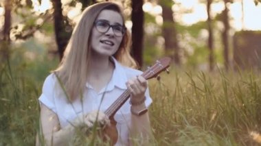 Ukulele çalan genç ve güzel bir kız. Gitar çalan beyaz bir kız. Genç güzel sarışın kadın gitar çalıyor ve çimlerin üzerinde otururken şarkı söylüyor. Kapat..