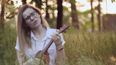 Ukulele çalan genç ve güzel bir kız. Gitar çalan beyaz bir kız. Genç güzel sarışın kadın gitar çalıyor ve çimlerin üzerinde otururken şarkı söylüyor. Kapat..
