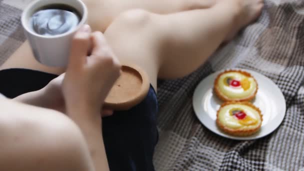 女の子はベッドで朝のコーヒーとケーキで目を覚ます 居心地の良い青いローブでベッドでコーヒーを飲む美しい若い女性 — ストック動画