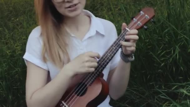 年轻美丽的女孩在弹奏四弦琴 白人女孩弹吉他 年轻美丽的金发女人坐在草地上弹奏四弦琴 靠近点 — 图库视频影像
