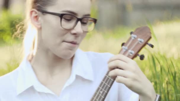 年轻美丽的女孩在弹奏四弦琴 白人女孩弹吉他 年轻美丽的金发女人坐在草地上弹奏四弦琴 靠近点 — 图库视频影像